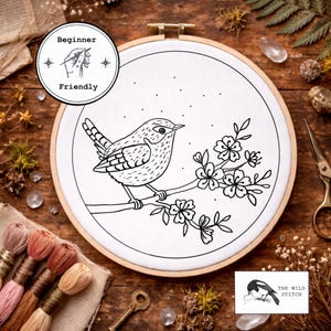 Könnte beinhalten: Ein schwarz-weißer Stickrahmen mit einem Vogel auf einem blühenden Zweig. Der Rahmen ist mit "Beginner Friendly" und "The Wild Stitch" beschriftet. Der Rahmen liegt auf einem rustikalen Holzgrund, umgeben von Stickgarn und Zubehör.