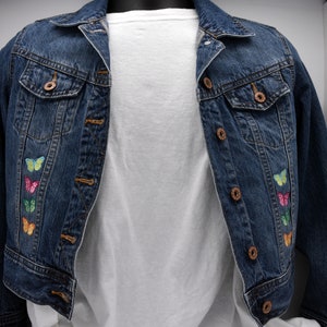 May include: A dark blue denim jacket with embroidered butterfly designs in various colors. The jacket has a classic button-up front, two chest pockets, and is paired with a white t-shirt underneath.