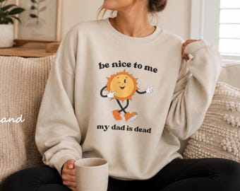 Könnte beinhalten: Sandfarbener Pullover mit dem Text "be nice to me my dad is dead" und einer Cartoon-Sonne. Die Sonne hat ein lächelndes Gesicht, Arme, Beine und orangefarbene Schuhe. Der Pullover ist ein lässiger, bequemer Stil.