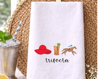 May include: White hand towel with a red hat, cocktail, and horse racing graphic. The word "trifecta" is printed below the graphics. The towel is hanging on a gold hook against a woven background.