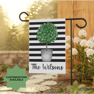 May include: A white garden flag with black stripes features a watercolor illustration of a green topiary tree in a gray pot. The text "The Wilsons" is printed in black at the bottom of the flag.