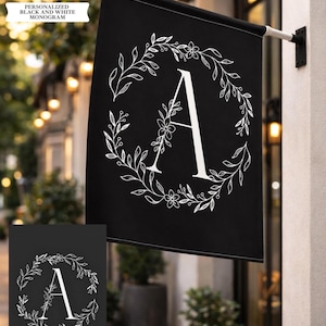 May include: Black flag with a white floral wreath design and the letter "A" in the center. The flag is hanging outside on a building. The text "PERSONALIZED BLACK AND WHITE MONOGRAM" is in a white banner in the upper left corner.
