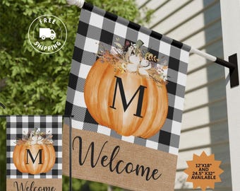 May include: A black and white checked welcome flag featuring a pumpkin with the letter "M" in the centre. The pumpkin is decorated with autumnal flowers and leaves. The bottom of the flag has a hessian texture with the word "Welcome" written in black.
