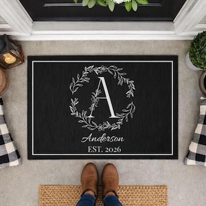 May include: Black rectangular doormat with a white border. The mat features a white floral wreath surrounding the letter "A", with the name "Anderson" and the year "EST. 2026" in a script font. The mat is placed in front of a door.