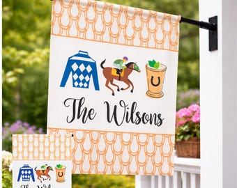 May include: A rectangular garden flag featuring an orange horseshoe pattern border, a blue jockey shirt, a brown horse with a rider, and a mint julep cup. The text reads The Wilsons in black script on a white background.