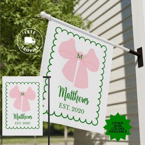 May include: A white garden flag with a pink bow design and the initial "M". The flag has a green scalloped border and the name "Matthews EST. 2020" in green script. A smaller version of the flag is also visible.
