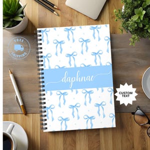 May include: A blue and white spiral bound notebook with a light blue ribbon pattern and the name "daphne" written in cursive on the front cover.