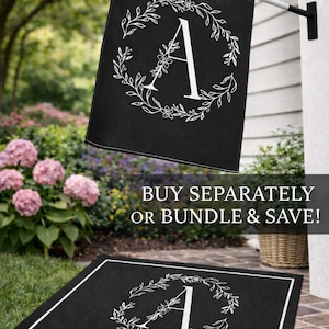 May include: Black outdoor flag and doormat set featuring a white floral wreath design around the letter "A". The flag is hanging on a white pole. The doormat is rectangular with a white border. The background includes greenery and pink flowers.