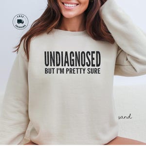 May include: A sand-colored sweatshirt with the text "UNDIAGNOSED BUT I'M PRETTY SURE" in black. The sweatshirt has long sleeves and a crew neck. The image also includes a "FREE SHIPPING" graphic.