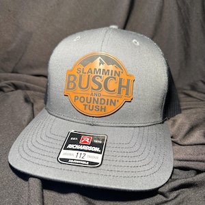 May include: Gray trucker hat with a black mesh back and a curved brim. The hat features a patch with the words "SLAMMIN' BUSCH AND POUNDIN' TUSH" in orange and white. A Richardson 112 Trucker tag is visible.