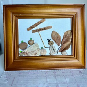 May include: A framed artwork featuring a 'Pumpkin Patch' sign made of wood. The artwork includes pebble pumpkins, a figure, and dried leaves. The frame is a warm brown color, and the background is a light canvas.
