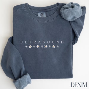 Peut inclure: Un sweat-shirt bleu foncé avec le texte "ULTRASOUND" en lettres blanches et une rangée de marguerites blanches avec des centres jaunes sous le texte. Le sweat-shirt est plié et posé à plat sur une surface blanche. Le mot "DENIM" est imprimé en lettres blanches dans le coin inférieur droit de l'image.