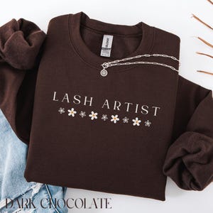 Peut inclure: Un sweat-shirt marron foncé avec le texte "LASH ARTIST" en blanc et des motifs de marguerites. Un collier argenté avec un pendentif soleil est drapé sur le sweat-shirt. Le sweat-shirt est plié, avec un jean visible.