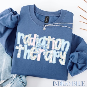 May include: Indigo blue sweatshirt with the words "Radiation Therapy" in a decorative font. The text is a mix of floral and striped patterns with a light blue bow. A silver necklace with a sun pendant is on the sweatshirt.