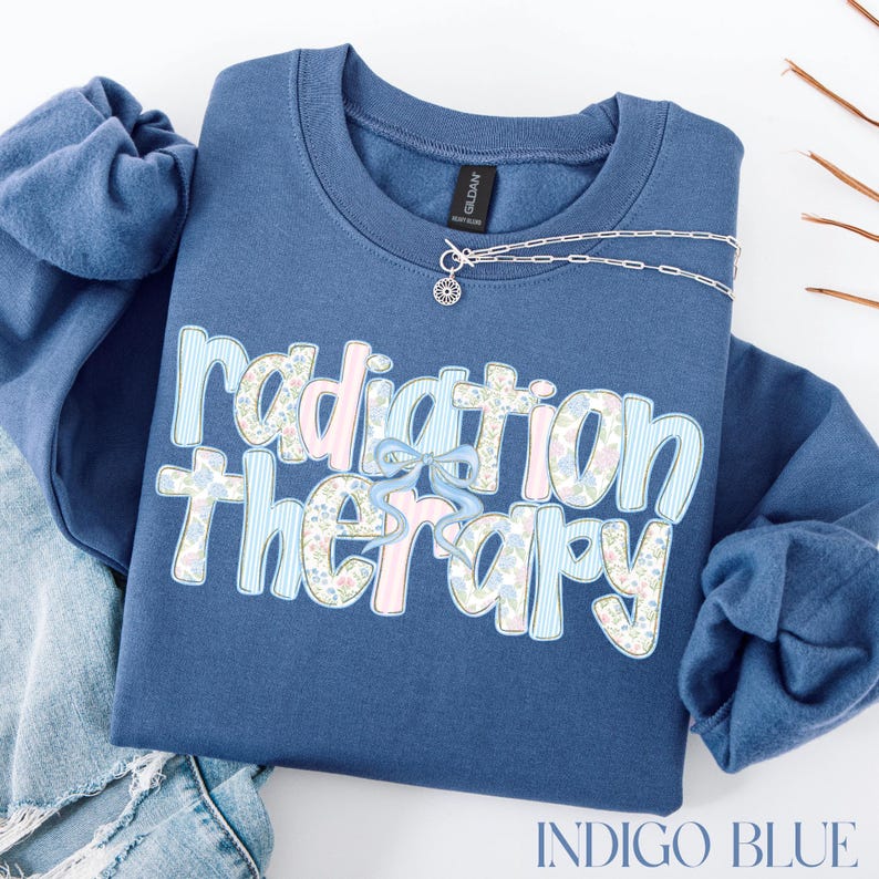 May include: Indigo blue sweatshirt with the words "Radiation Therapy" in a decorative font. The text is a mix of floral and striped patterns with a light blue bow. A silver necklace with a sun pendant is on the sweatshirt.