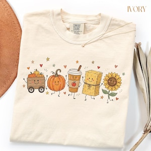 May include: An ivory t-shirt featuring a graphic print of a wooden wagon with pumpkins, a large pumpkin, a coffee cup with an apple, a hay bale, and a sunflower, all with smiling faces. The shirt has a Comfort Colors brand label at the neck.