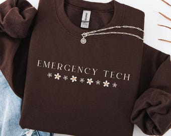 May include: A dark brown sweatshirt with the text "EMERGENCY TECH" in white lettering and a row of white daisy flowers below the text. The sweatshirt is made of a soft, comfortable material and has a relaxed fit.
