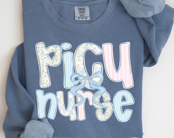May include: Blue jean colored sweatshirt with the words "PICU nurse" in a decorative font. The letters are decorated with floral patterns, pink stripes, and a blue bow. The sleeves are rolled up, and the Comfort Colors tag is visible.