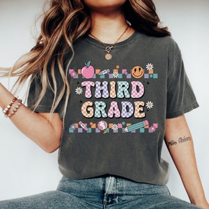 May include: A dark gray t-shirt with a colorful graphic design that says "Third Grade" in a retro font. The graphic includes a checkered pattern, an apple, a crayon, a smiley face, a pencil, a globe, and flowers.