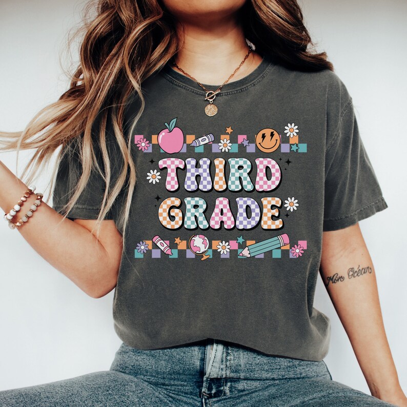 May include: A dark gray t-shirt with a colorful graphic design that says "Third Grade" in a retro font. The graphic includes a checkered pattern, an apple, a crayon, a smiley face, a pencil, a globe, and flowers.
