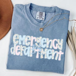 May include: A blue Comfort Colors t-shirt with the words "Emergency Department" in a floral and striped design. A gold necklace with a pendant is draped across the shirt.