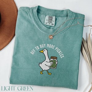 May include: Light green Comfort Colors t-shirt with the phrase "OFF TO BUY MORE PICKLES" and a cartoon goose holding a jar of pickles. The shirt also has a small tag and a silver necklace.