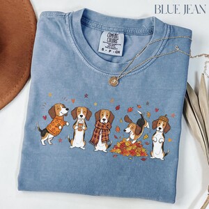 Puede incluir: Una camiseta azul de cuello redondo con un gr&aacute;fico de cinco perros beagle con ropa de oto&ntilde;o, incluyendo jers&eacute;is, bufandas y un sombrero de bellota, rodeados de hojas que caen y peque&ntilde;os corazones. La camiseta tiene una etiqueta en el cuello.
