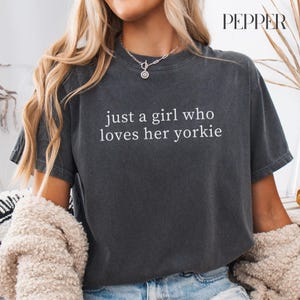 May include: Dark gray t-shirt with the text "just a girl who loves her yorkie" in white. The shirt is a crew neck style and has short sleeves. The model is wearing a necklace and a cream-colored sweater.