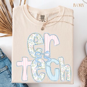 May include: Ivory-colored t-shirt with the text "ER TECH" in a floral and striped design. The letters are outlined in pink and blue, with a blue bow. A gold necklace with a pendant is draped on the shirt.