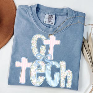 May include: A blue Comfort Colors t-shirt with the text "GT TECH" in a floral and striped design. The letters are outlined in pink and blue, with a light blue bow. A gold necklace is draped across the shirt.