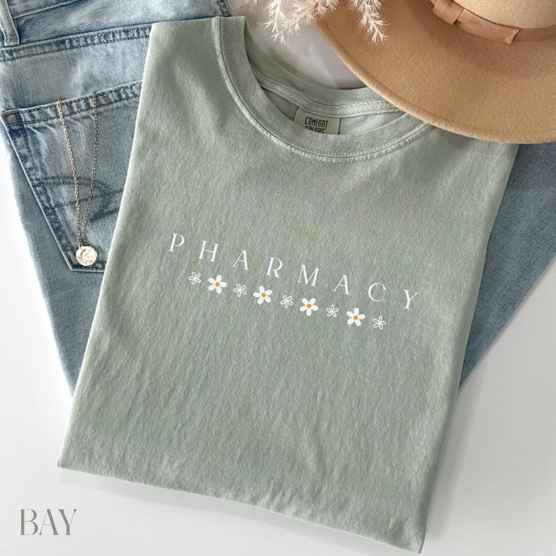 May include: A light green t-shirt with the word "PHARMACY" printed in white with small white flowers underneath. The shirt is laying on a pair of blue jeans.