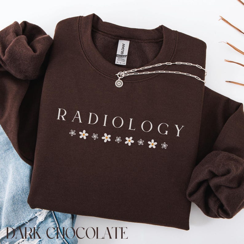 May include: A dark brown sweatshirt with the text "RADIOLOGY" in white lettering and a row of white daisy flowers below the text. The sweatshirt is made of a soft, comfortable material and has a relaxed fit.
