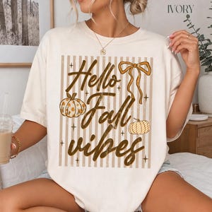 May include: An ivory short-sleeve t-shirt featuring a graphic print with vertical tan stripes, two pumpkins with polka dot patterns, a brown ribbon bow, and the text Hello Fall vibes in a brown script font, accented by small four-pointed stars.