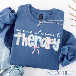 May include: Indigo blue sweatshirt with the words "occupational therapy" in a decorative font. The word "therapy" is in pink and blue floral print. A silver necklace with a sunburst pendant is on the sweatshirt.