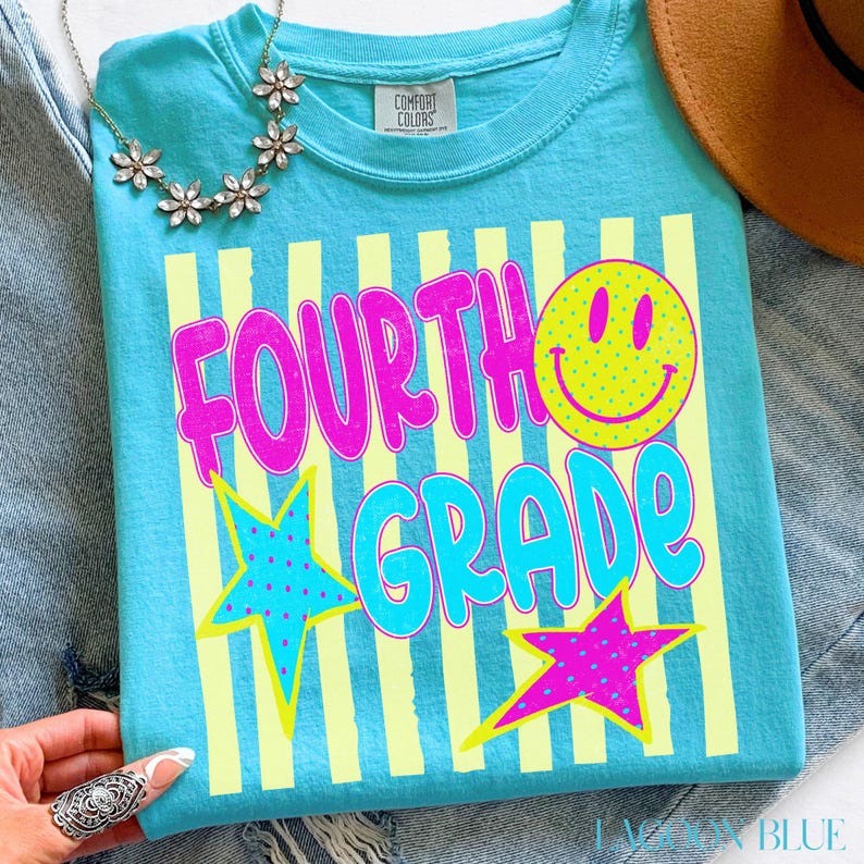 Op de afbeelding: Een Lagoon Blue t-shirt met de woorden "FOURTH GRADE" in roze en turquoise, een gele smiley en twee sterren. Het shirt heeft gele verticale strepen en de woorden "COMFORT COLORS" op het label.