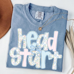May include: A blue Comfort Colors t-shirt with the words "head start" in a floral and striped design. A gold necklace with a pendant is draped across the shirt. The shirt is folded and has a small tag.