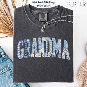 May include: A dark gray t-shirt with the word "GRANDMA" in a patchwork design. The letters are a mix of blue gingham, floral, and solid white fabrics. A silver necklace with a flower pendant rests on the shirt. The text "Not Real Stitching Print Only" is at the top.