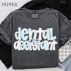 Op de afbeelding: Een donkergrijs lang mouwshirt met de woorden "dental assistant" in een speels lettertype. De letters zijn versierd met bloemen- en streepmotieven in pastelroze en -blauw, met een lichtblauwe strik.