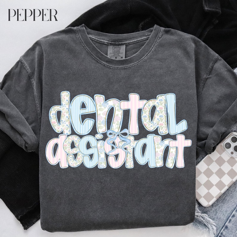Op de afbeelding: Een donkergrijs lang mouwshirt met de woorden "dental assistant" in een speels lettertype. De letters zijn versierd met bloemen- en streepmotieven in pastelroze en -blauw, met een lichtblauwe strik.