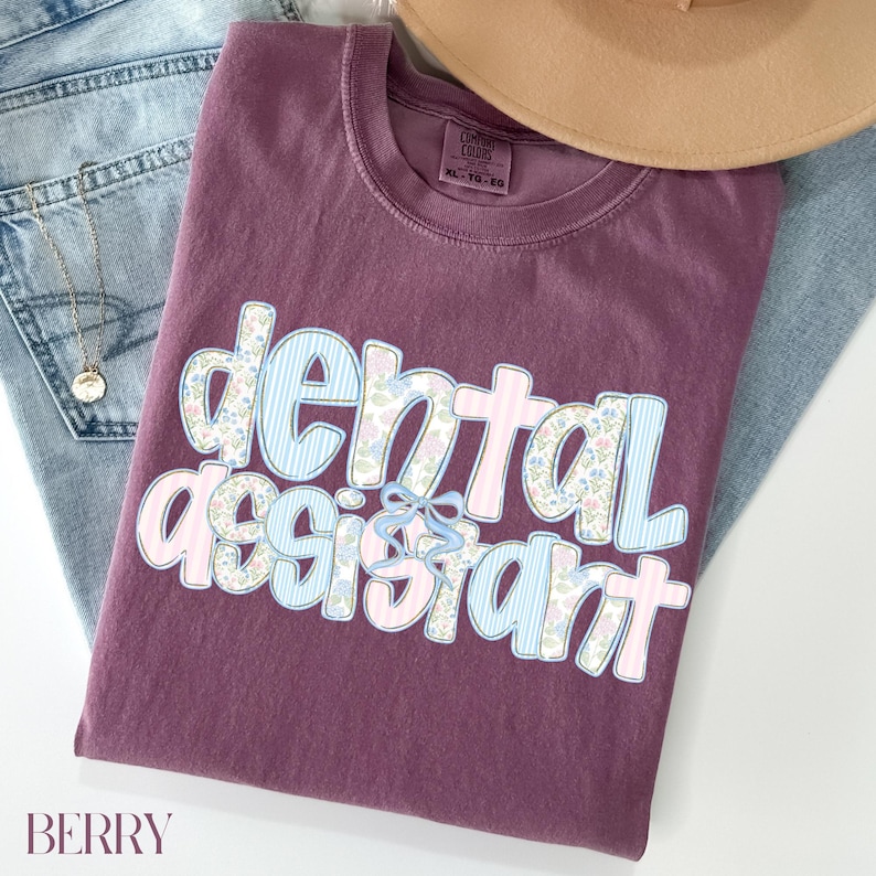 Op de afbeelding: Een bessenkleurig T-shirt met de woorden "dental assistant" in een grillig ontwerp. De tekst bevat een combinatie van bloemen- en streepjespatronen in lichtblauw en roze, met een kleine blauwe strik. Het shirt ligt op een spijkerbroek.