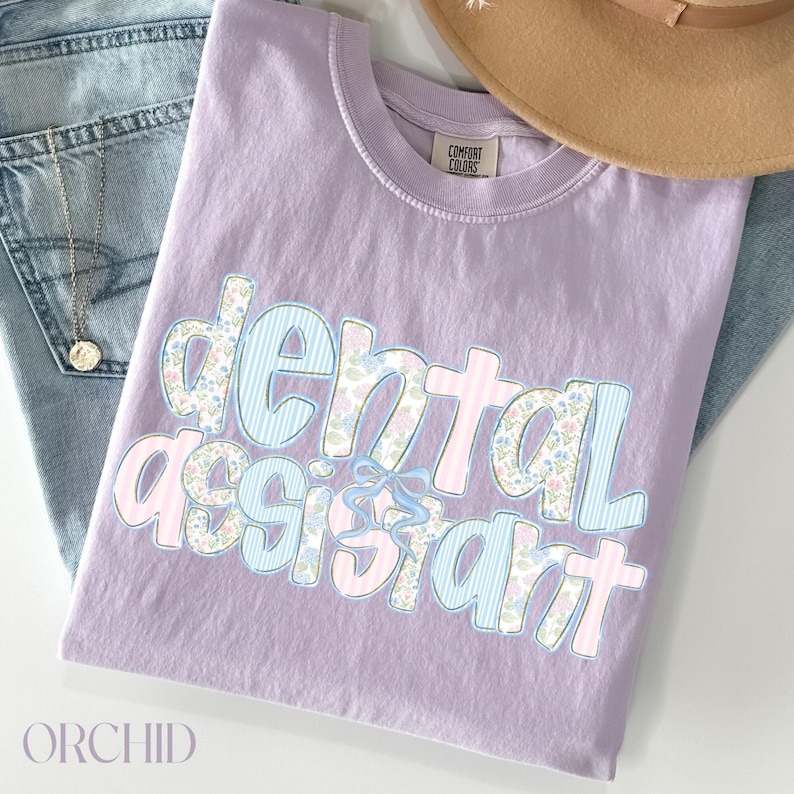Op de afbeelding: Een lila T-shirt met de woorden "dental assistant" in een decoratief lettertype. De letters zijn een mix van bloemen- en streepmotieven in lichtblauw en roze. Het shirt ligt op een spijkerbroek en een beige hoed.
