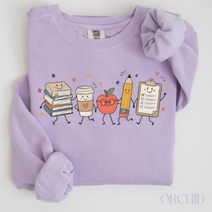 Op de afbeelding: Een lichtpaarse sweater met ronde hals en een grafische print van een stapel boeken, een koffiebeker, een appel met een bril, een potlood en een klembord, allemaal met lachende gezichten. Het Comfort Colors label is zichtbaar in de nek.