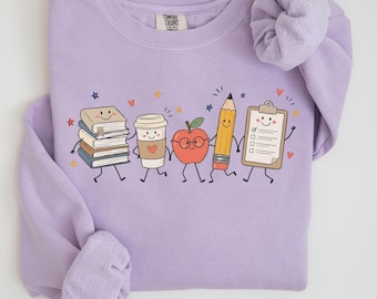 Può includere: Una felpa girocollo color viola chiaro con una stampa grafica raffigurante una pila di libri, una tazza di caffè, una mela con gli occhiali, una matita e una cartellina, tutti con facce sorridenti. L'etichetta Comfort Colors è visibile sul collo.