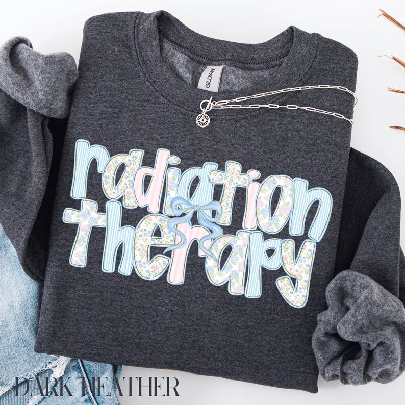 May include: Dark heather gray sweatshirt with the words "Radiation Therapy" in a floral and striped design. A silver necklace with a sun pendant is draped across the top. The sweatshirt is folded on a white surface.