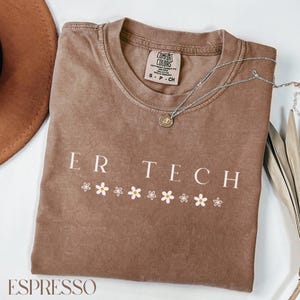 May include: A brown t-shirt with the text "ER TECH" in white lettering and a row of white daisies below. The shirt is folded and has a tag that says "COMFORT COLORS" on it.