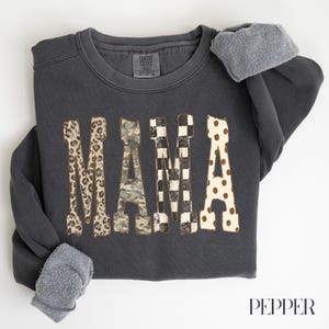 Op de afbeelding: Een antracietkleurige sweater met lange mouwen en een fleecevoering. Het woord MAMA staat in grote blokletters op de borst gedrukt, waarbij elke letter een ander patroon heeft: luipaardprint, camouflage, zwart-wit geblokt en stippen.