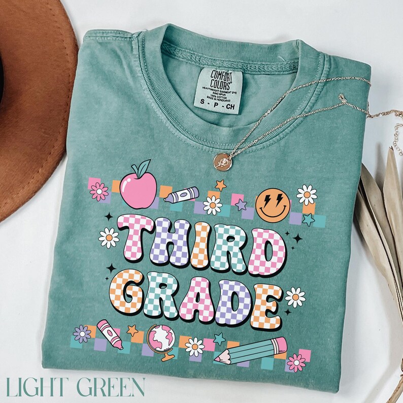 May include: Light green t-shirt with a checkered design and the words "Third Grade" in colorful letters. The shirt is decorated with school-themed graphics, including an apple, a pencil, a globe, and a smiley face.