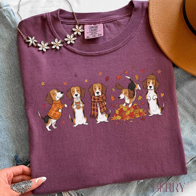 Puede incluir: Una camiseta color ciruela con un gr&aacute;fico de cinco perros beagle con atuendos y poses oto&ntilde;ales, incluyendo uno con su&eacute;ter de calabazas, uno sosteniendo una bebida, uno con bufanda de cuadros, uno jugando entre hojas y uno con sombrero de bellota.