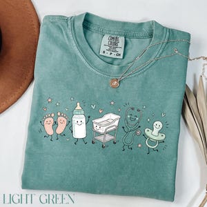 May include: A sage green short-sleeve t-shirt featuring a graphic print of cartoon baby items including feet, a bottle, a bassinet, a stethoscope, and a pacifier. The shirt has a Comfort Colors label at the neck and a size S tag.