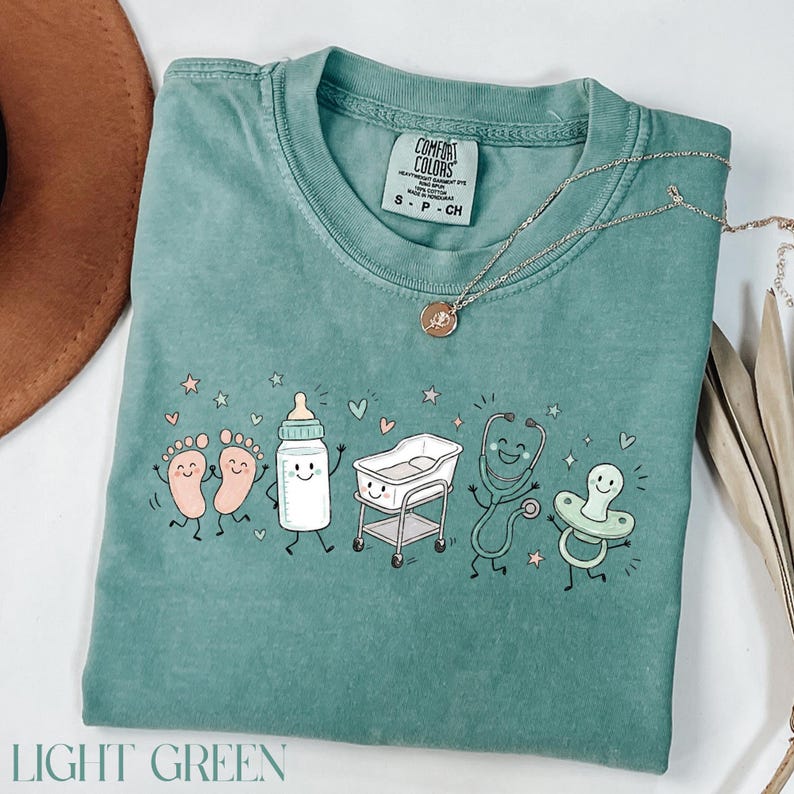 May include: A sage green short-sleeve t-shirt featuring a graphic print of cartoon baby items including feet, a bottle, a bassinet, a stethoscope, and a pacifier. The shirt has a Comfort Colors label at the neck and a size S tag.